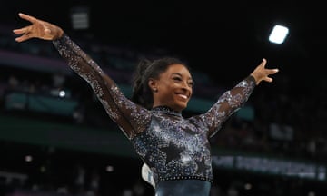 Biles stands, grinning, with her arms outstretched after her routine. She wears a shiny, metallic-effect blue, navy and black leotard. The top half of it has stars across the front. There are crystals across the top half of her leotard and on the arms