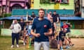 Freddie Flintoff smiles to camera while throwing a cricket ball from one hand to the other with men and boys behind him, some with cricket bats, in front of a brightly coloured wooden building