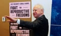 A man with white hair gestures his hand as he stands in front of signs saying 'Vice President Kamala Harris Fight For Reproductive Freedoms'.