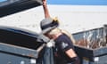 A blonde woman wearing a black T-shirt and a gray and black hat lifts up the lid to a dumpster and peers in