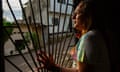 A trans woman looks out of a grill over a window on to a street