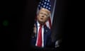A man in a suit-and-tie speaks on stage in front of an American flag