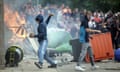 A young man with hood pulled up throws a brick, as another stands nearby. Flames and a crowd of people are behind