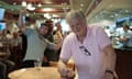 Tim Martin smiles for a photo while leaning on a table in a pub, while two young men photobomb him with gestures behind him