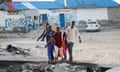 Five men carry a body in a red plastic sheet across the sand with a restaurant in the background
